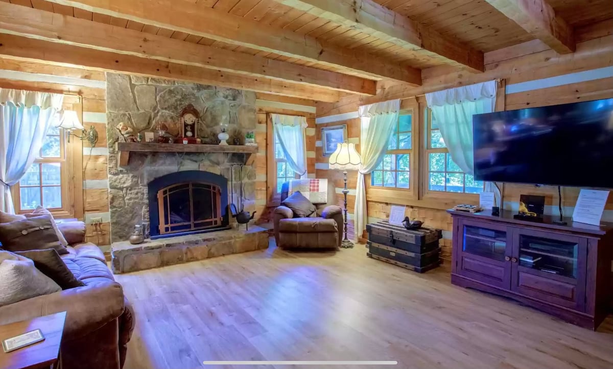 A cozy living room is characterized by wooden beams and a stone fireplace, providing a rustic feel. Soft lighting from table lamps and natural light through several windows creates a warm ambiance. Comfortable seating includes a sofa and armchair, enhancing relaxation.