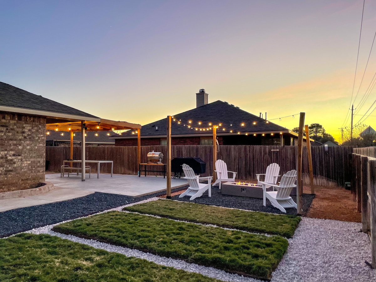 An outdoor space is presented with string lights illuminating a covered area. A dining table is positioned beside a grill, while seating is arranged around a firepit. Green grass contrasts with gravel pathways, and a warm sunset visible in the background adds to the inviting setting.