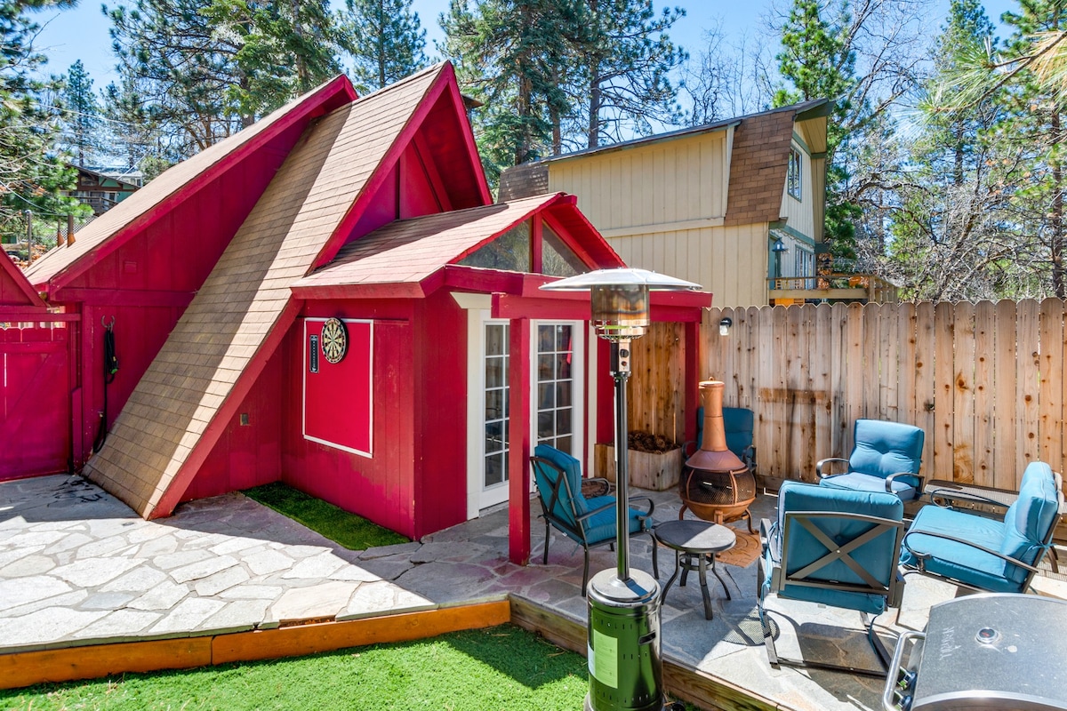 A vibrant red cabin is depicted in a private outdoor space, featuring a flagstone patio and a green turf area. Comfortable blue chairs are arranged around a patio fire feature, while a decorative wall clock adds a touch of charm. Nearby, a fenced-in area provides privacy.