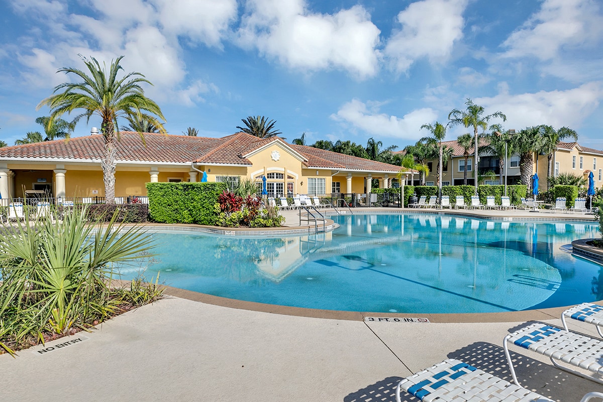 The resort features a large outdoor pool surrounded by neatly landscaped greenery and tropical palm trees. Sun loungers are arranged along the pool's edge. The clubhouse sits adjacent to the pool, offering a welcoming atmosphere for relaxation.
