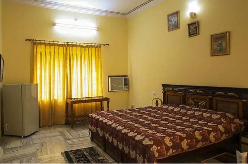 A spacious room features a large bed draped in a patterned coverlet. The walls are painted in soft yellow, accented by framed artwork. Natural light filters through floor-length golden curtains, illuminating a wooden table and an air conditioning unit in the corner.