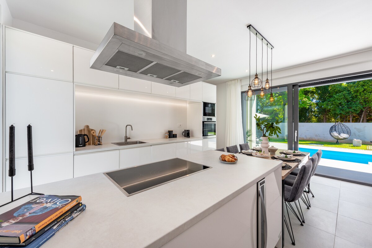 A modern kitchen is presented with sleek white cabinetry and a spacious island. Under-cabinet lighting illuminates the countertop, while pendant lights hang above. A dining area is visible, featuring several chairs surrounding a table, with glass doors leading to a pool area surrounded by greenery.