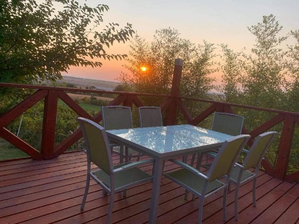 A wooden terrace features a rectangular dining table surrounded by six chairs. The scene is enhanced by the sunset illuminating the horizon, framed by trees in the background.