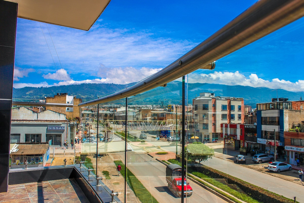 A scenic view of the surrounding neighborhood is captured through a glass railing. The image showcases a wide street lined with various buildings and shops, with distant mountains visible under a clear blue sky.