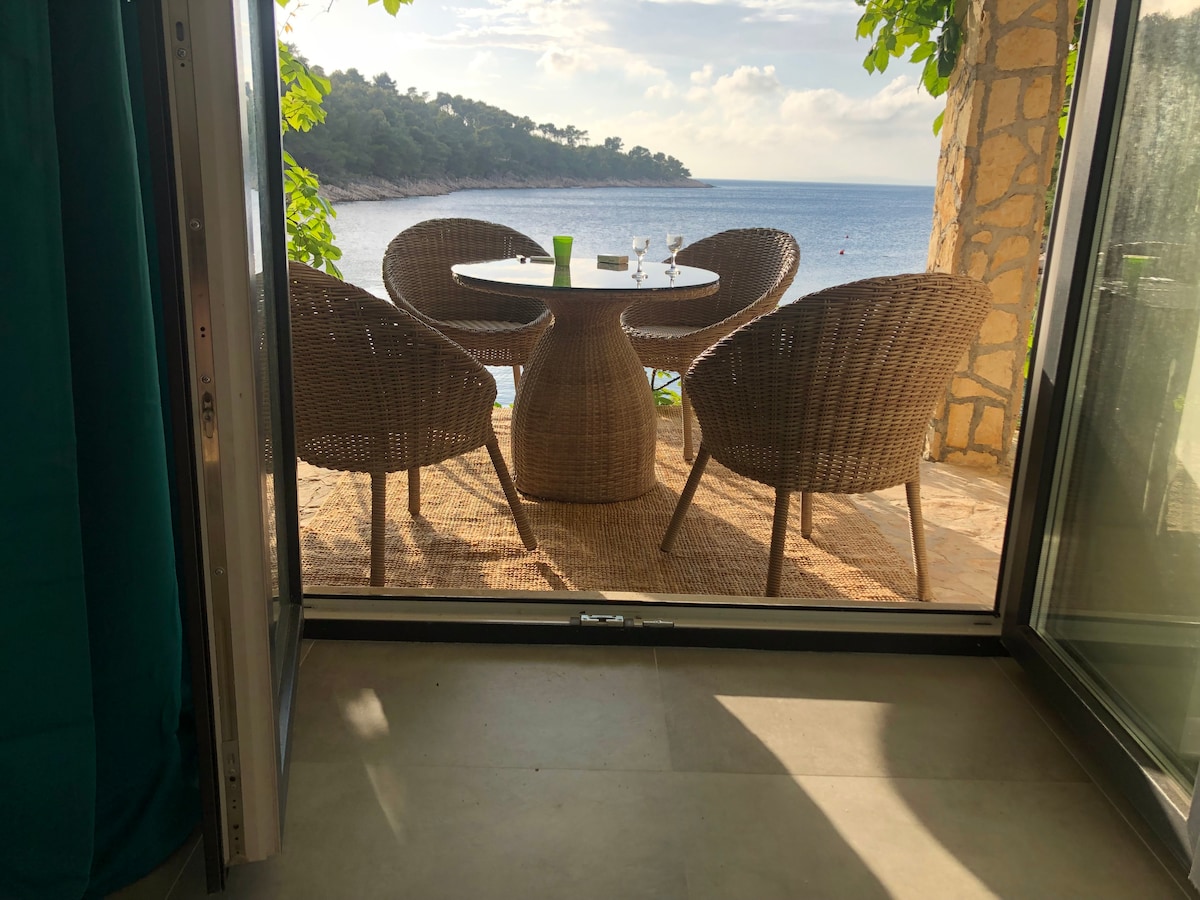 A view is presented from inside the house, showcasing a covered outdoor dining area with a round table and three woven chairs. The setting overlooks the calm sea and lush greenery, inviting relaxation and enjoyment of the natural surroundings.