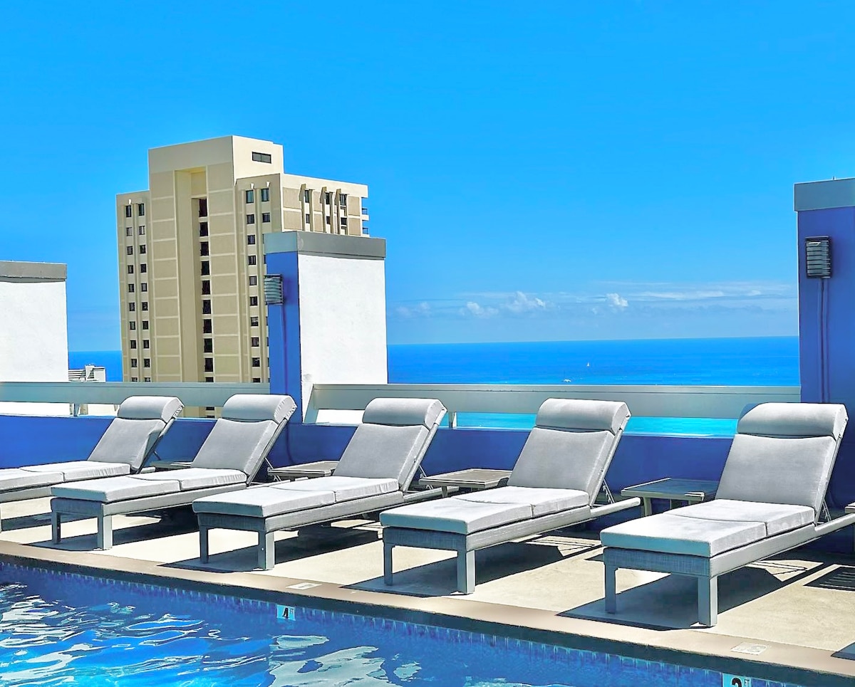 A rooftop pool area showcases several lounge chairs arranged along the edge, inviting relaxation. The swimming pool reflects the clear blue sky, with distant ocean views visible beyond the ledge. Nearby buildings provide a modern backdrop against the serene landscape.