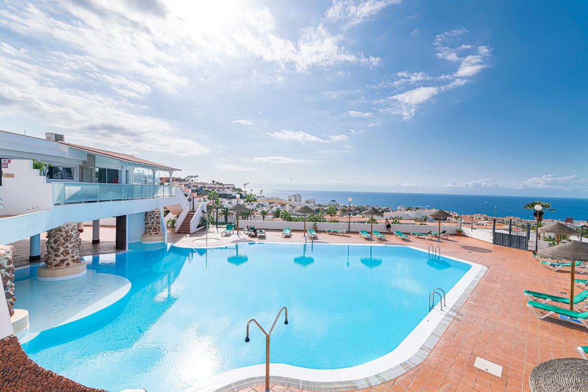 A large swimming pool is shown, surrounded by sun loungers and umbrellas. The area offers expansive views of the ocean and distant mountains under a bright sky. The modern architecture integrates seamlessly with the inviting pool area, providing a tranquil setting for relaxation.