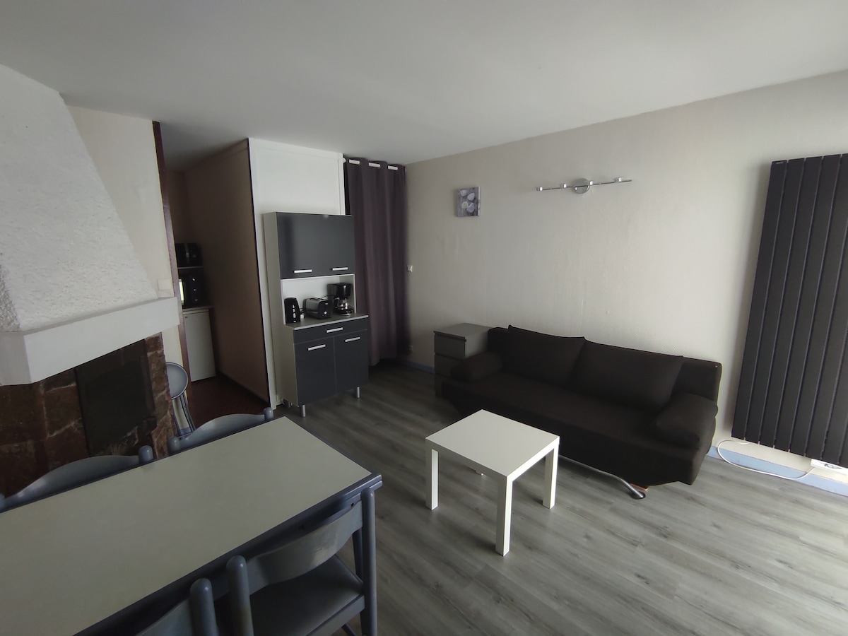 A casual living space includes a dark sofa and a light coffee table, contrasted by grey flooring. A kitchenette is present, equipped with modern appliances. Two walls are partially covered by curtains, and a simple wall art piece enhances the overall decor.