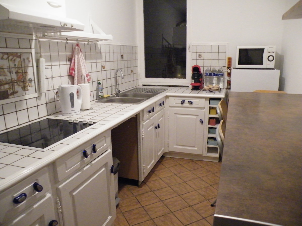 The kitchen features white cabinetry with blue handles, tiled countertops, and a mix of modern and traditional appliances. A sink and stovetop are included, alongside a small countertop. A microwave and coffee machine sit on the counter, providing essential cooking amenities.
