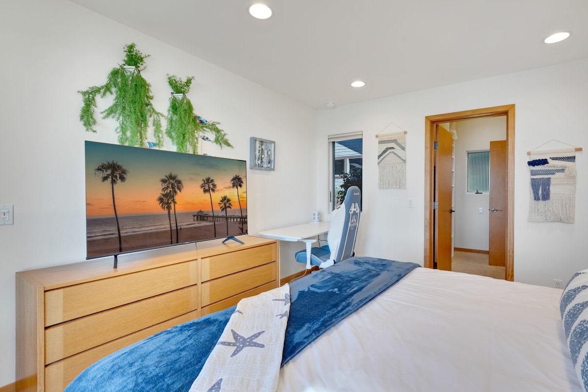 A cozy bedroom features a spacious bed adorned with soft linens and a decorative throw. A large television stands on a wooden dresser, displaying a sunset beach scene. A work desk and chair are positioned beside a window that lets in natural light.