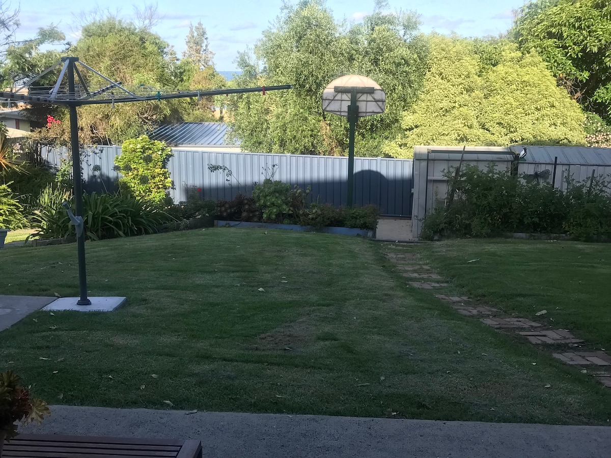 A grassy outdoor space features neatly maintained landscaping, including a paved pathway leading toward a garden area. A clothesline stands prominently on the left, while a shade structure adds a functional element. Lush greenery surrounds the area, providing a sense of privacy.