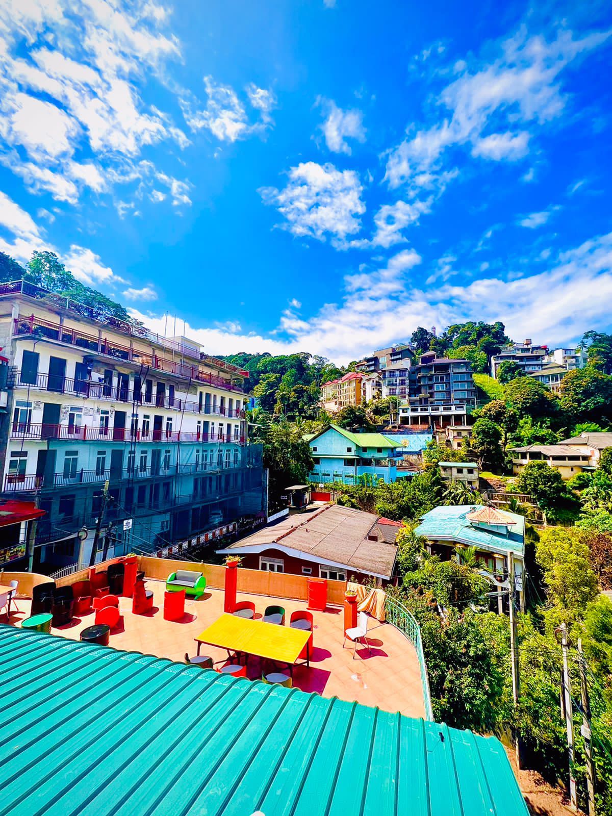 The image captures a vibrant outdoor terrace with colorful furniture, including tables and chairs. Surrounding hills and buildings are visible under a bright blue sky scattered with clouds, providing a lively urban view enhanced by greenery.