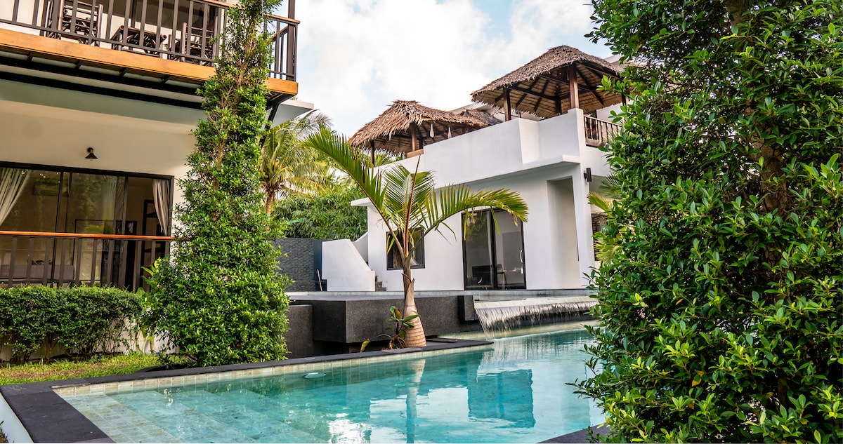 A modern apartment complex is surrounded by lush tropical gardens, featuring a serene communal pool with a gentle waterfall. The pool reflects clear blue water, while neighboring areas are edged with vibrant greenery and shaded structures. Comfortable lounging spots are visible for relaxation.