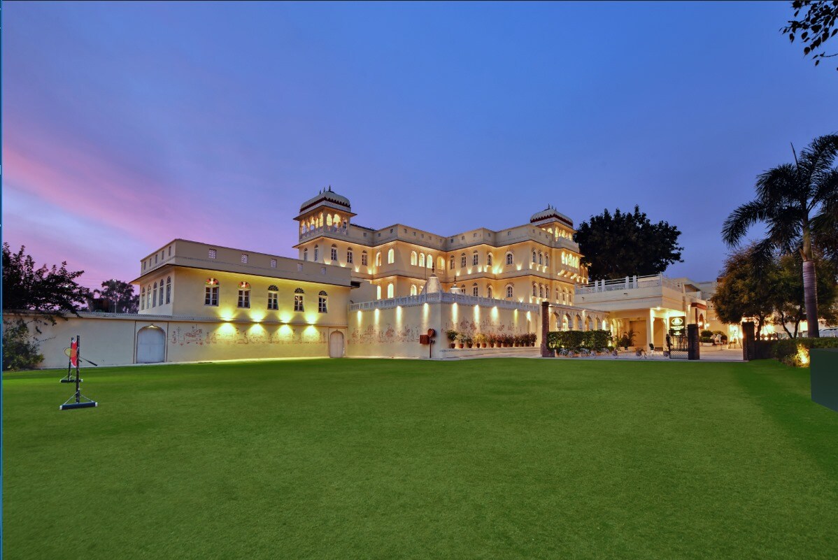 The majestic structure of Jagjit Mahal is highlighted against a twilight sky, showcasing its architectural grandeur. Lush green lawns surround the building, and soft outdoor lighting enhances the elegant ambiance, inviting guests to explore the spacious surroundings.
