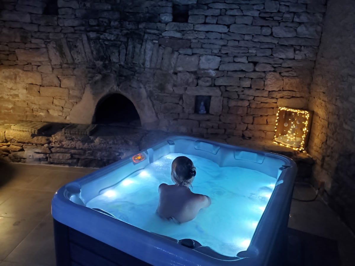 A private jacuzzi is seen, featuring soft blue lighting that enhances the tranquil ambiance. The setting includes a stone wall and a rustic fireplace, contributing to a cozy atmosphere. A person is immersed in the water, offering a sense of relaxation.