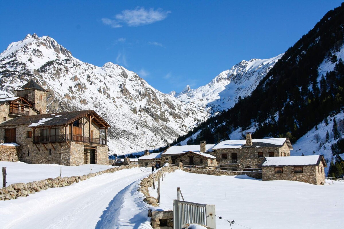 A scenic winter landscape is presented, featuring rustic stone buildings surrounded by a blanket of snow. Towering mountains rise in the background, complemented by a clear blue sky with a few scattered clouds. A snow-covered path leads through the tranquil setting.