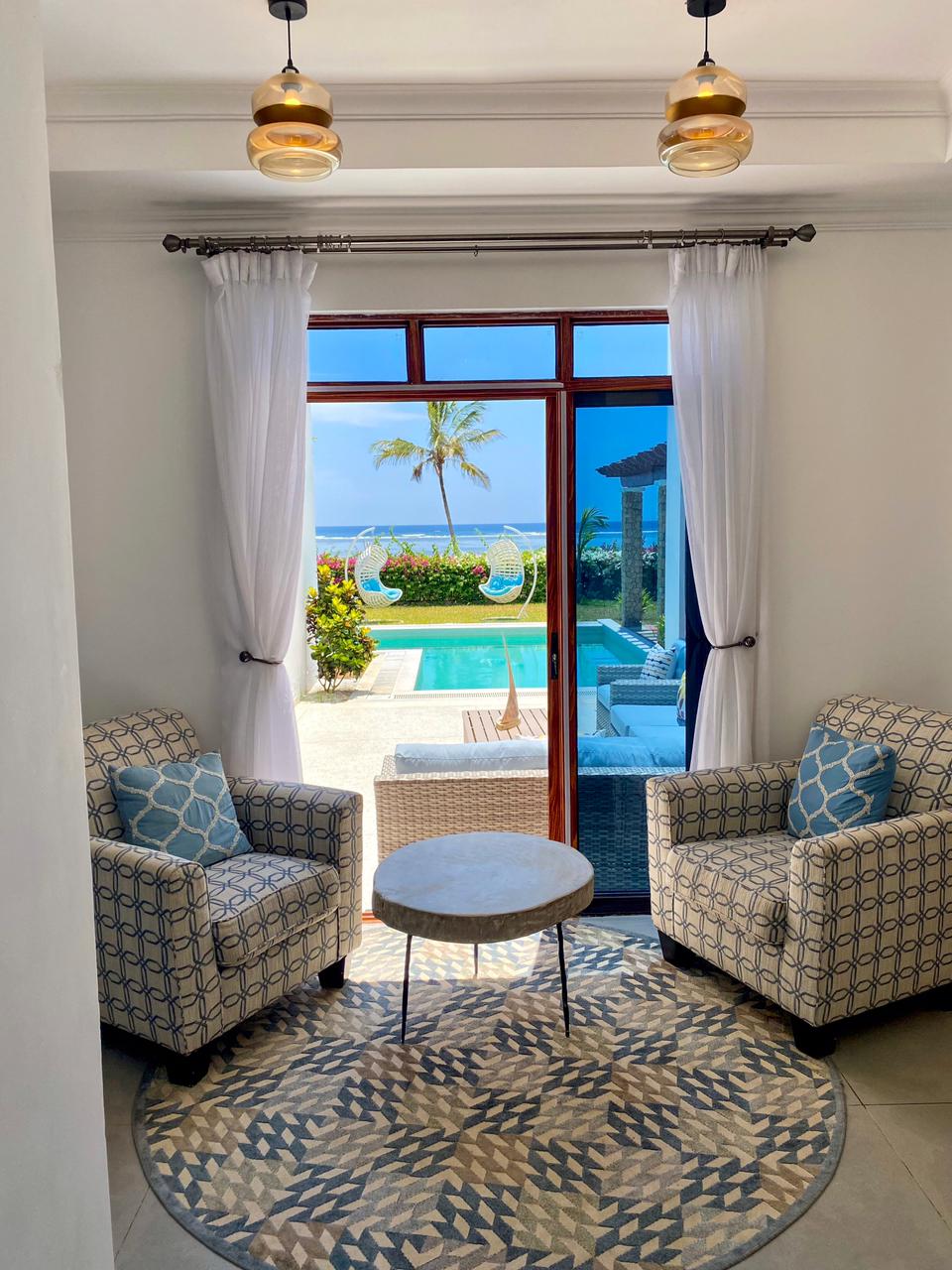 Two patterned armchairs are positioned near a circular table, with natural light filtering in through large glass doors. The view reveals a pool surrounded by tropical greenery, leading to the sparkling expanse of the ocean.