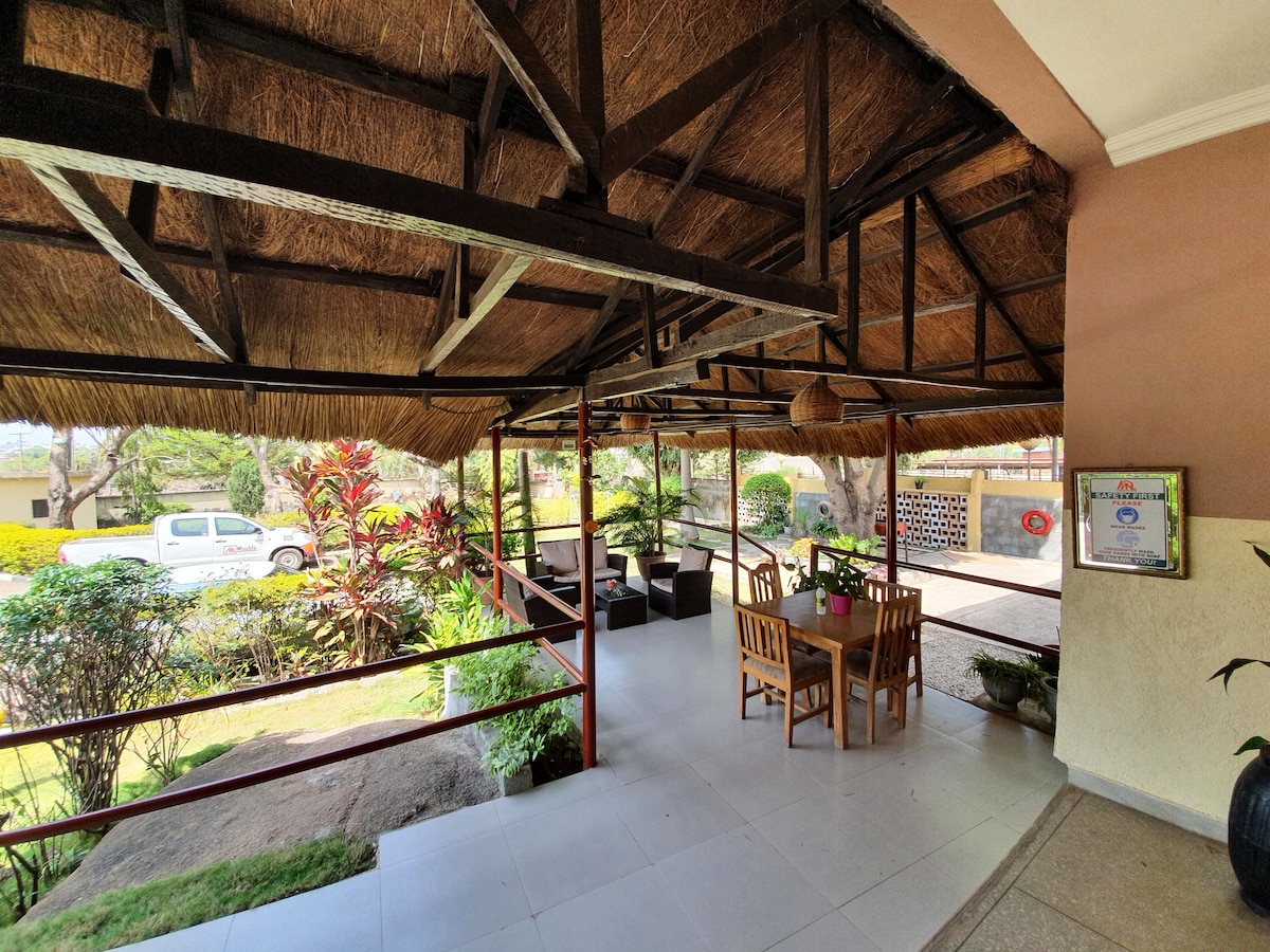 An inviting outdoor seating area features wooden tables and chairs beneath a thatched roof. Lush greenery surrounds the space, with potted plants and garden foliage visible. The open design creates a connection to the outdoors, providing a relaxed environment for guests.