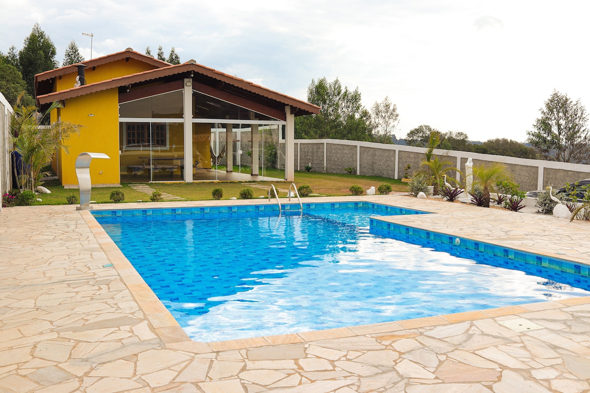 A bright outdoor area is highlighted by a large swimming pool, featuring clear blue water. Surrounding the pool, a spacious deck is made of stone tiles, with lush greenery and decorative plants enhancing the landscape. A large glass exterior facilitates views into the living spaces.