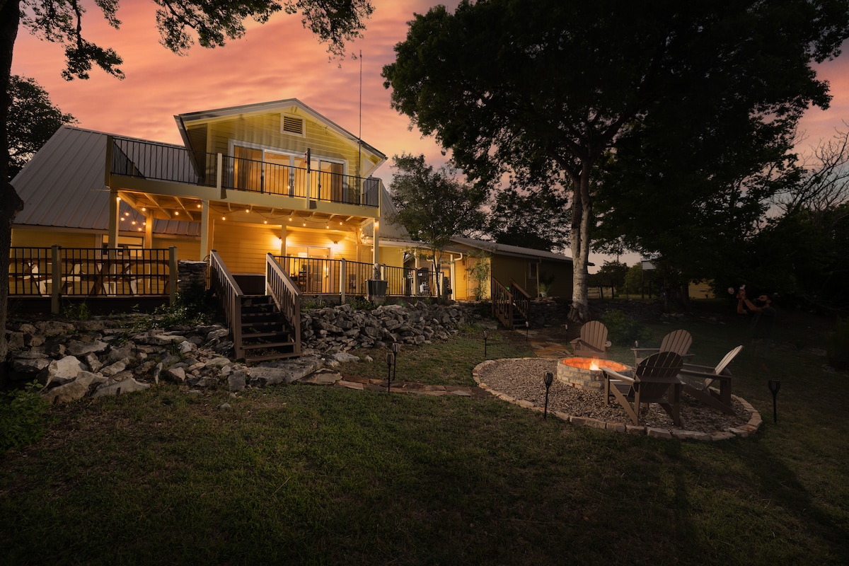 The exterior of a two-story country home is illuminated as dusk approaches, showcasing warm lighting. A comfortable firepit area, surrounded by stone, features four seating chairs. The lush lawn and nearby trees complement the serene setting, providing a peaceful retreat.