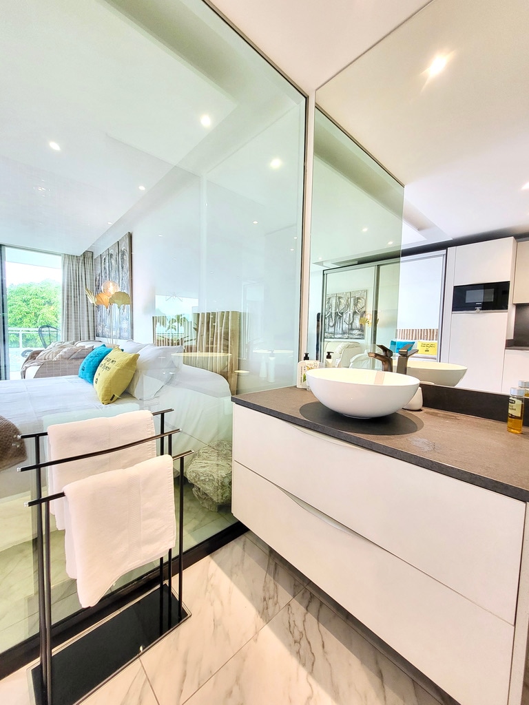 A modern bathroom features a sleek glass partition separating it from the sleeping area. A minimalist white sink is complemented by a stylish countertop, while plush towels hang neatly on a holder. Natural light enhances the bright, airy feel of the space.