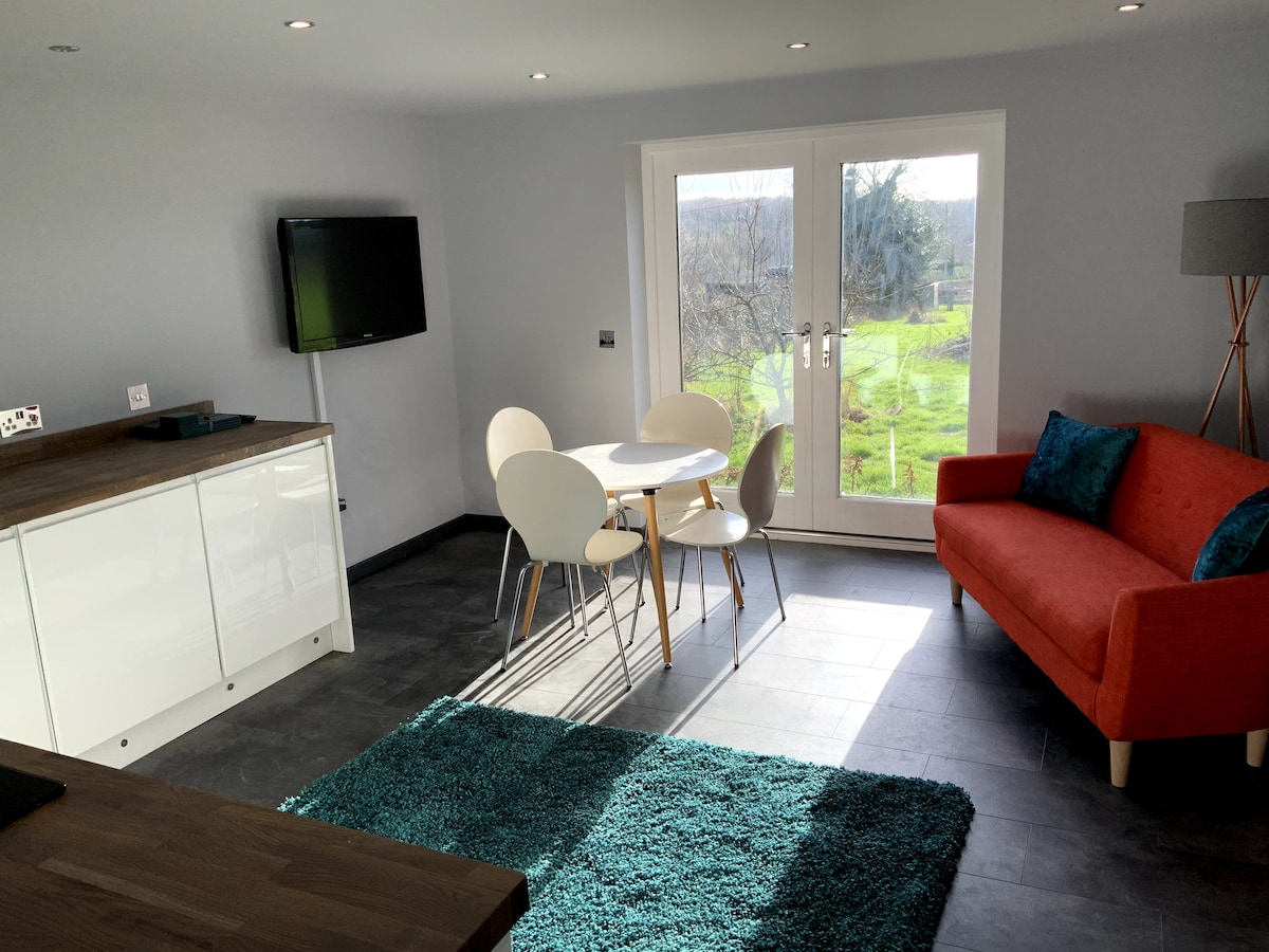 A spacious living area features a bright orange sofa and a circular dining table with four white chairs. A flat-screen TV is mounted on the wall, and large glass doors lead to a view of the surrounding countryside. Natural light floods the room, highlighting the blue area rug.