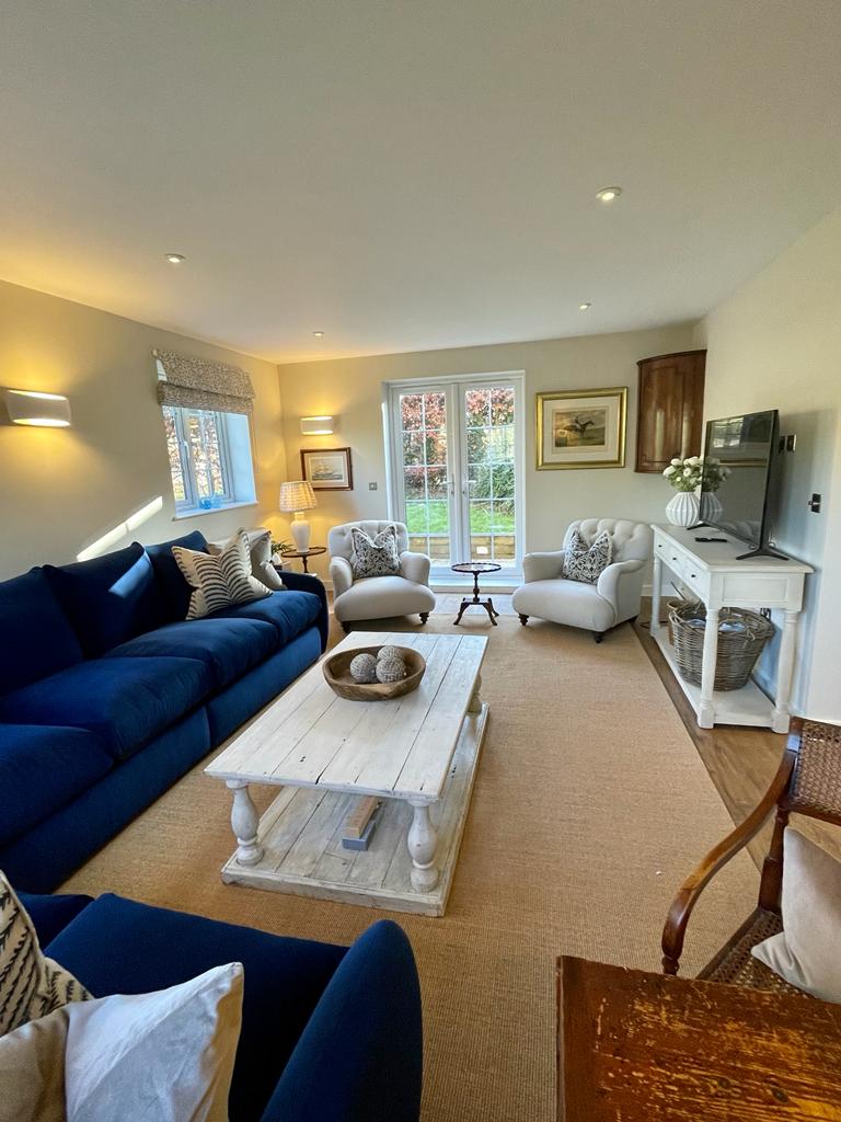 A sitting area is arranged with a deep blue sofa and two accent chairs, creating a comfortable space. A wooden coffee table displays decorative bowls, while a flat-screen television is positioned against the wall. Natural light enters through glass doors, leading to the garden.