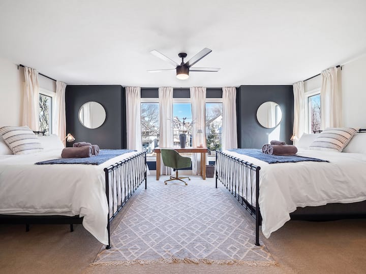 Chumba cha kulala angavu na chenye nafasi kubwa chenye vitanda viwili pacha, fremu nyeusi za kisasa, madirisha makubwa na eneo la dawati la kati lililojaa mwanga wa asili.
