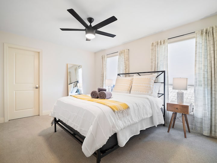 Chumba cha kulala angavu kilicho na fremu ya kisasa ya kitanda cheusi, matandiko safi meupe yenye rangi ya manjano na madirisha makubwa yanayoingiza mwanga wa asili.
