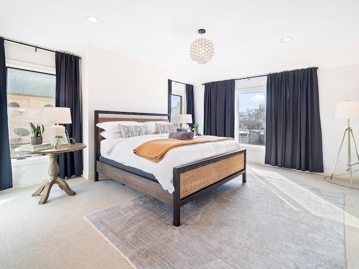 Chumba cha kulala angavu na chenye nafasi kubwa chenye madirisha makubwa, fanicha za kisasa na kitanda chenye starehe kilicho na mapazia meusi.
