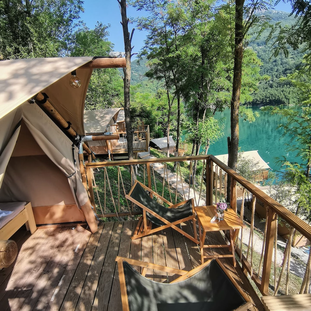 A private terrace features wooden deck chairs and a small table with a vase of flowers. The tranquil lake and lush greenery are visible, providing a natural setting. Glamping tents can be seen in the background, nestled among the trees.
