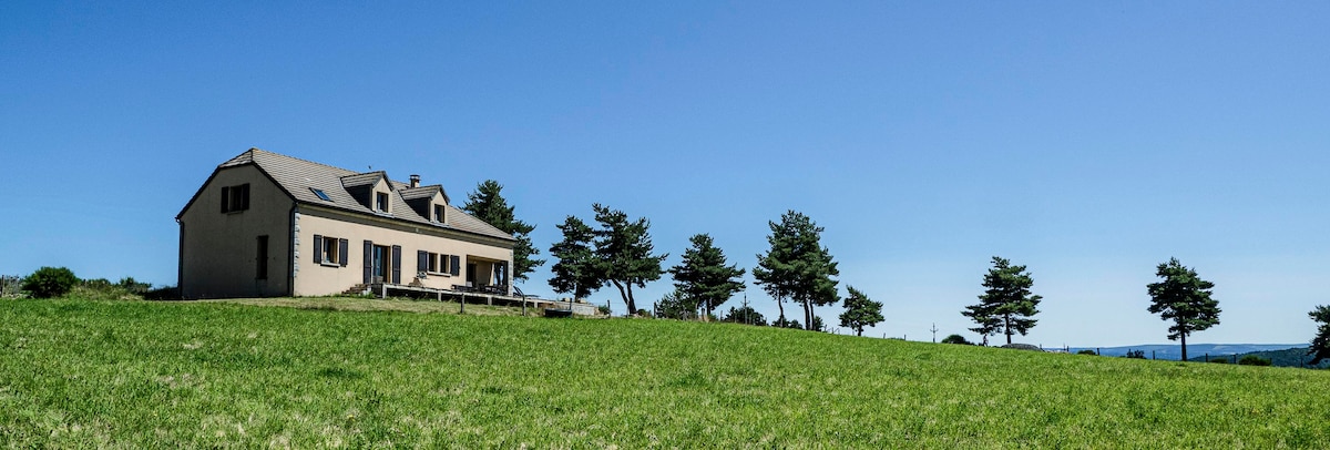 A spacious house is situated amidst a vast green landscape, with a clear blue sky overhead. Several trees provide a natural setting around the property, and large windows reflect the serene outdoor environment.