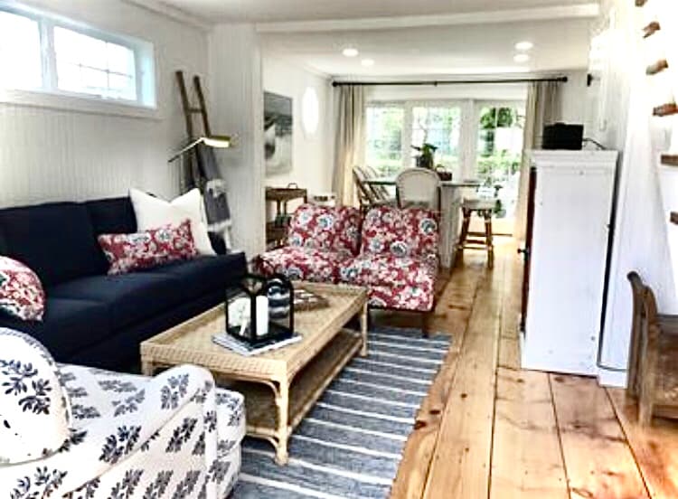 A cozy living area displays a dark blue sofa and two floral-patterned chairs, complemented by a wicker coffee table. Light wood flooring connects this space to the dining area, with natural light streaming through large windows that offer views of the outdoors.