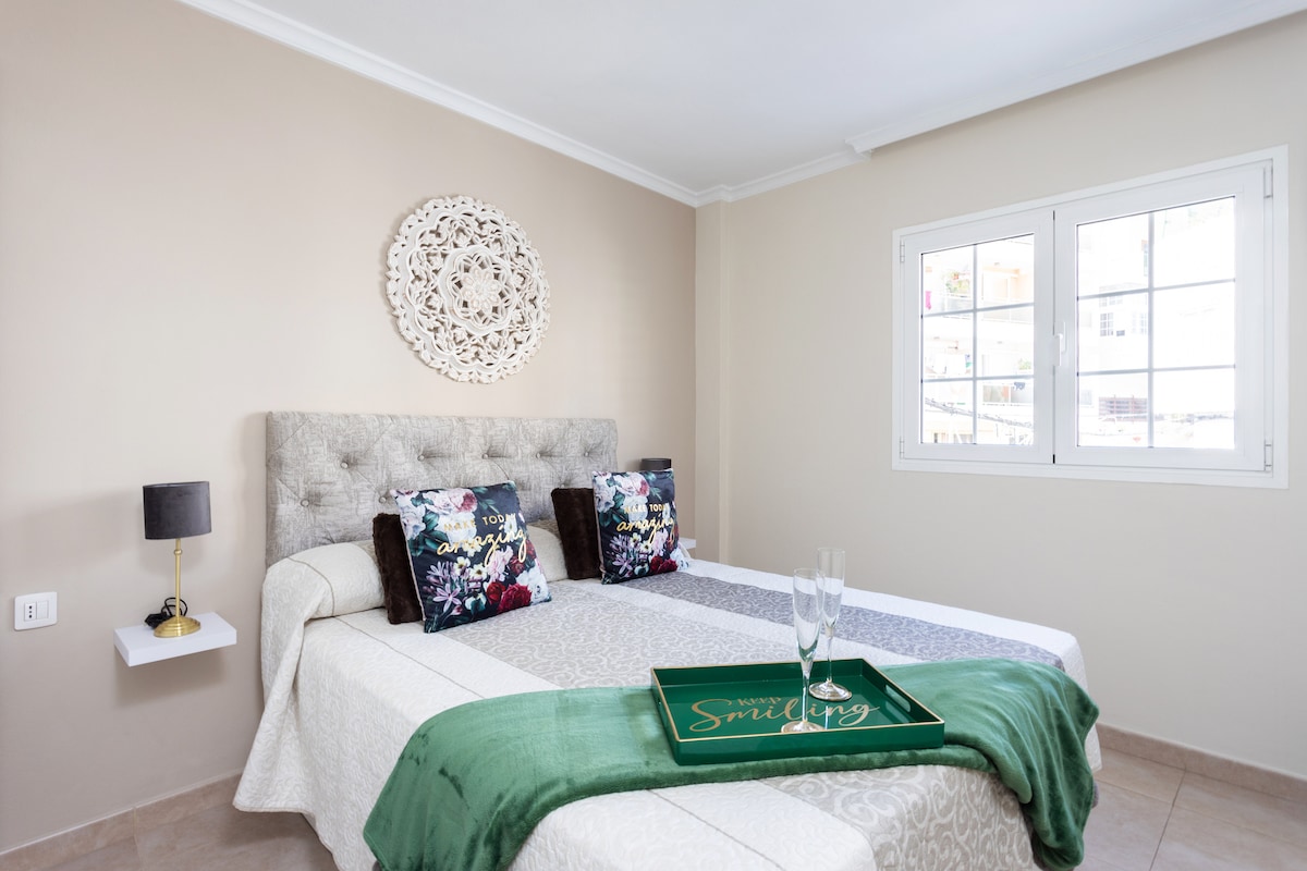 A well-lit bedroom features a spacious bed adorned with a decorative quilt. Two cushions with floral patterns are placed on the headboard. A tray with glasses is positioned at the foot of the bed, while natural light enters through multiple windows with white frames.