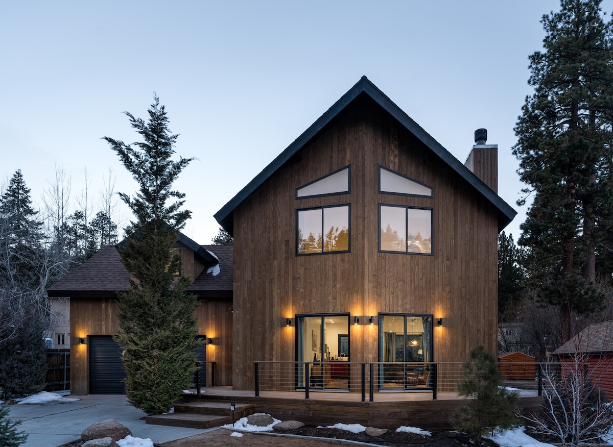 The exterior of a mid-century modern home is showcased, featuring a wooden facade and large windows. Soft lighting accentuates the architectural details. Snow patches are visible on the ground, and trees frame the property, contributing to a natural mountain setting.