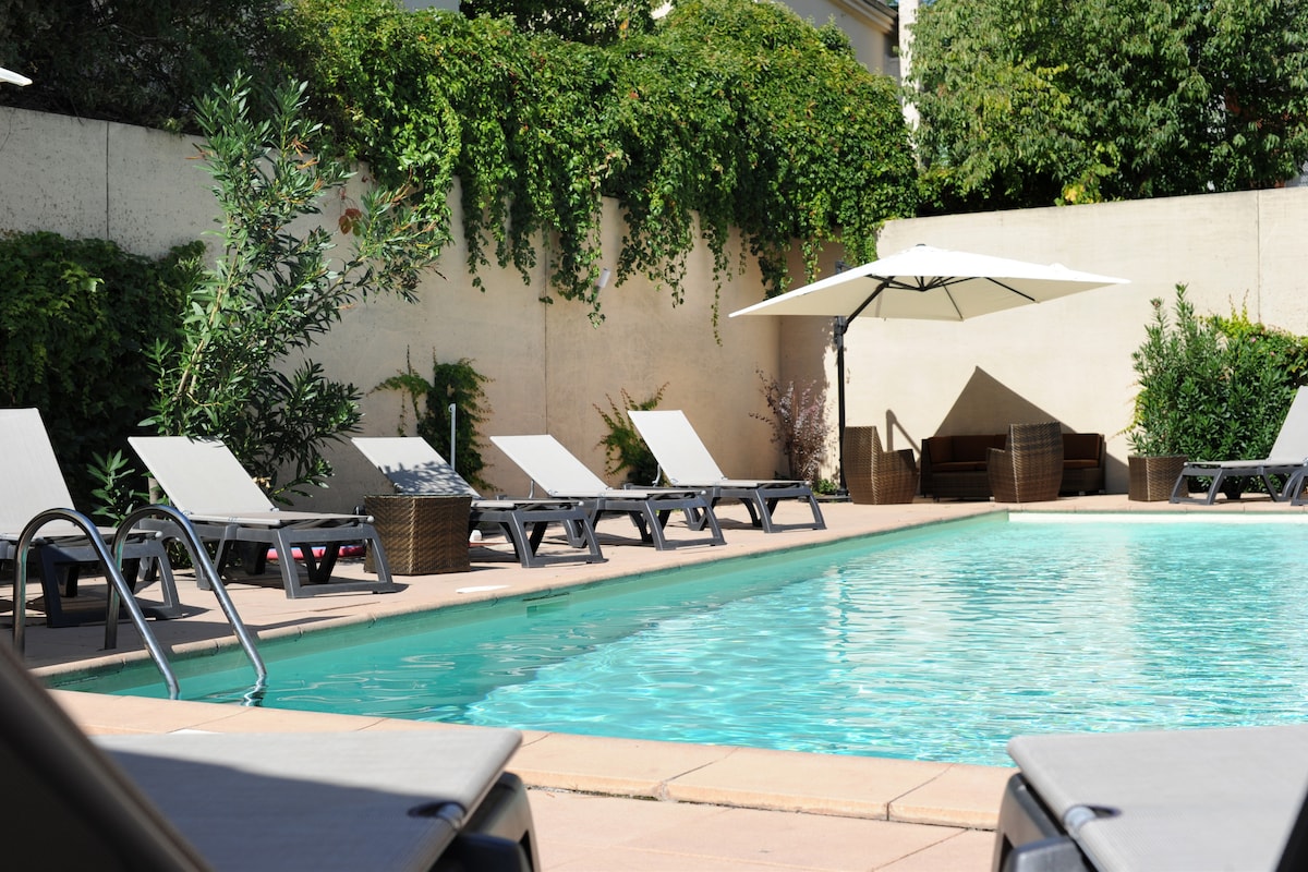 A swimming pool is surrounded by several lounge chairs, offering a relaxing setting. An umbrella provides shade over a seating area with wicker furniture situated near the pool. Lush greenery borders the space, enhancing the tranquil environment.