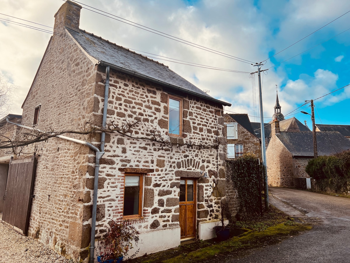 The cottage features a charming stone exterior with a wooden front door framed by wisteria. Two windows allow natural light to enter, and a sloped roof adds character. A gravel pathway leads from the street, with surrounding cottages and a church steeple visible in the background.