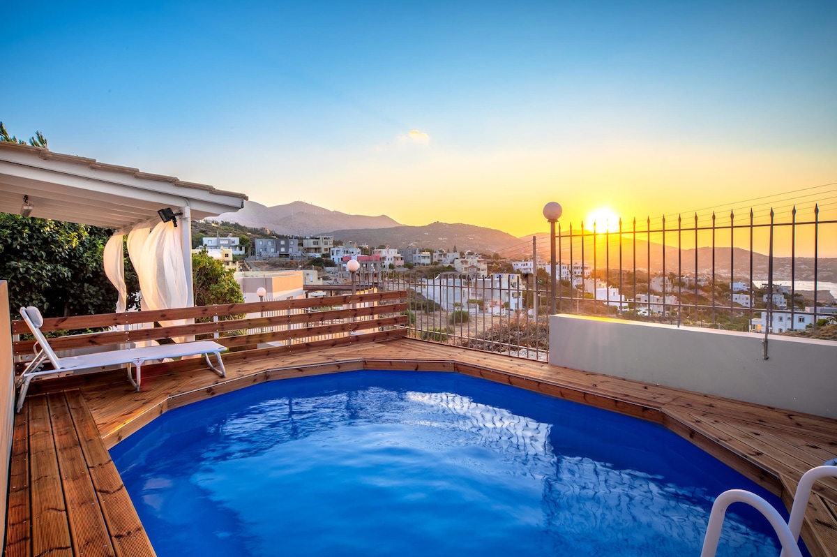 A swimming pool is framed by wooden decking, offering a serene view of the surrounding hillside and sunset. Lounge chairs are positioned nearby, creating a space for relaxation while enjoying the scenic landscape.