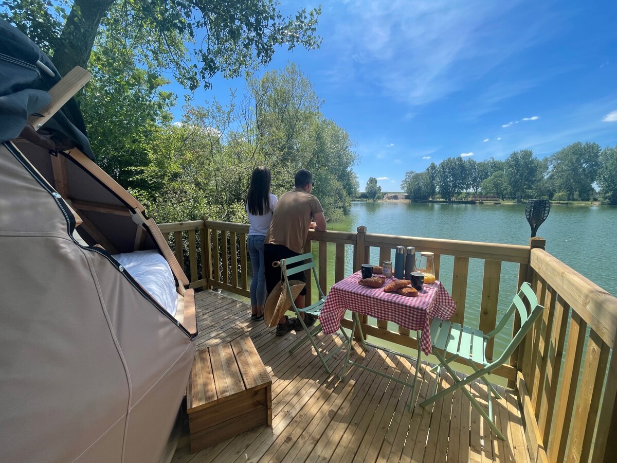 A wooden terrace overlooks a serene body of water, featuring a small table set with breakfast. Two individuals stand by the railing, enjoying the view. The setting is surrounded by greenery, providing a peaceful atmosphere.