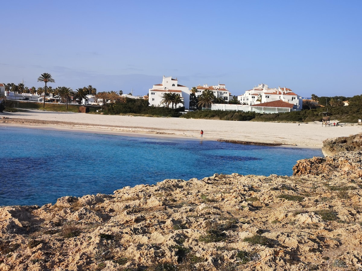 A shoreline view captures a gentle slope of sandy beach leading to calm blue waters. A cluster of white buildings is visible in the background, surrounded by greenery and palm trees. The rocky foreground adds texture to the scene, enhancing the natural beauty of the location.