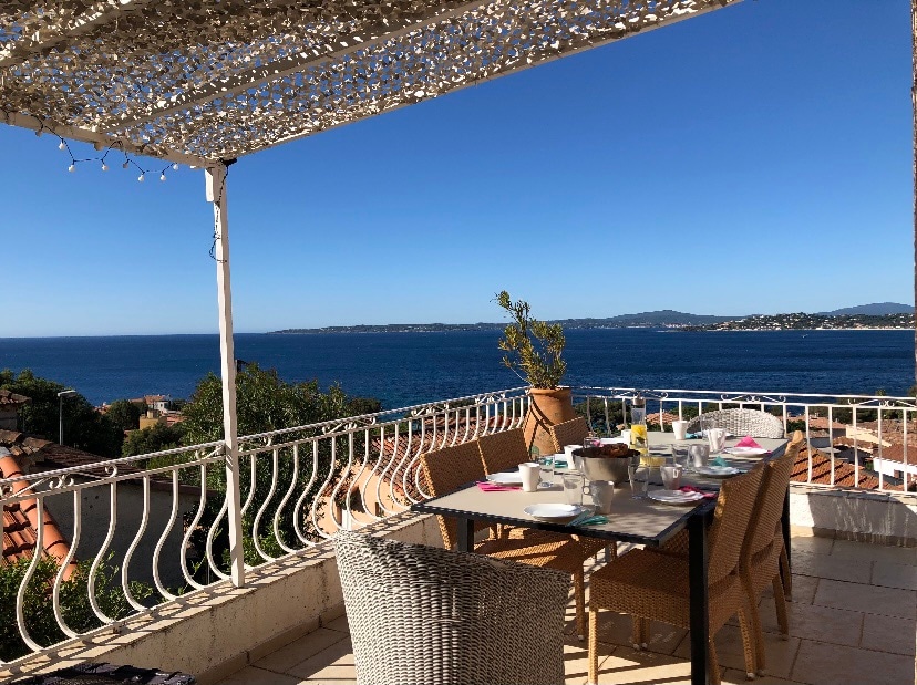 A terrace is depicted, featuring a dining table and woven chairs. Breakfast items are arranged on the table, with clear views of the sea and distant coastal hills in the background. A sheltered area above provides shade, enhancing the outdoor dining experience.