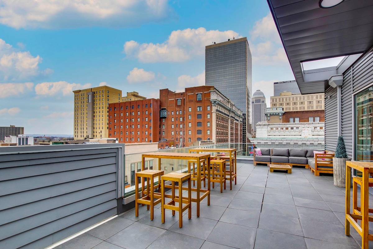 A spacious rooftop terrace is featured, equipped with wooden tables and benches arranged for relaxing views of the city skyline. The soft blue sky complements the urban landscape, showcasing a mix of modern and historic buildings in the background. A sense of openness is created by the surrounding railing.