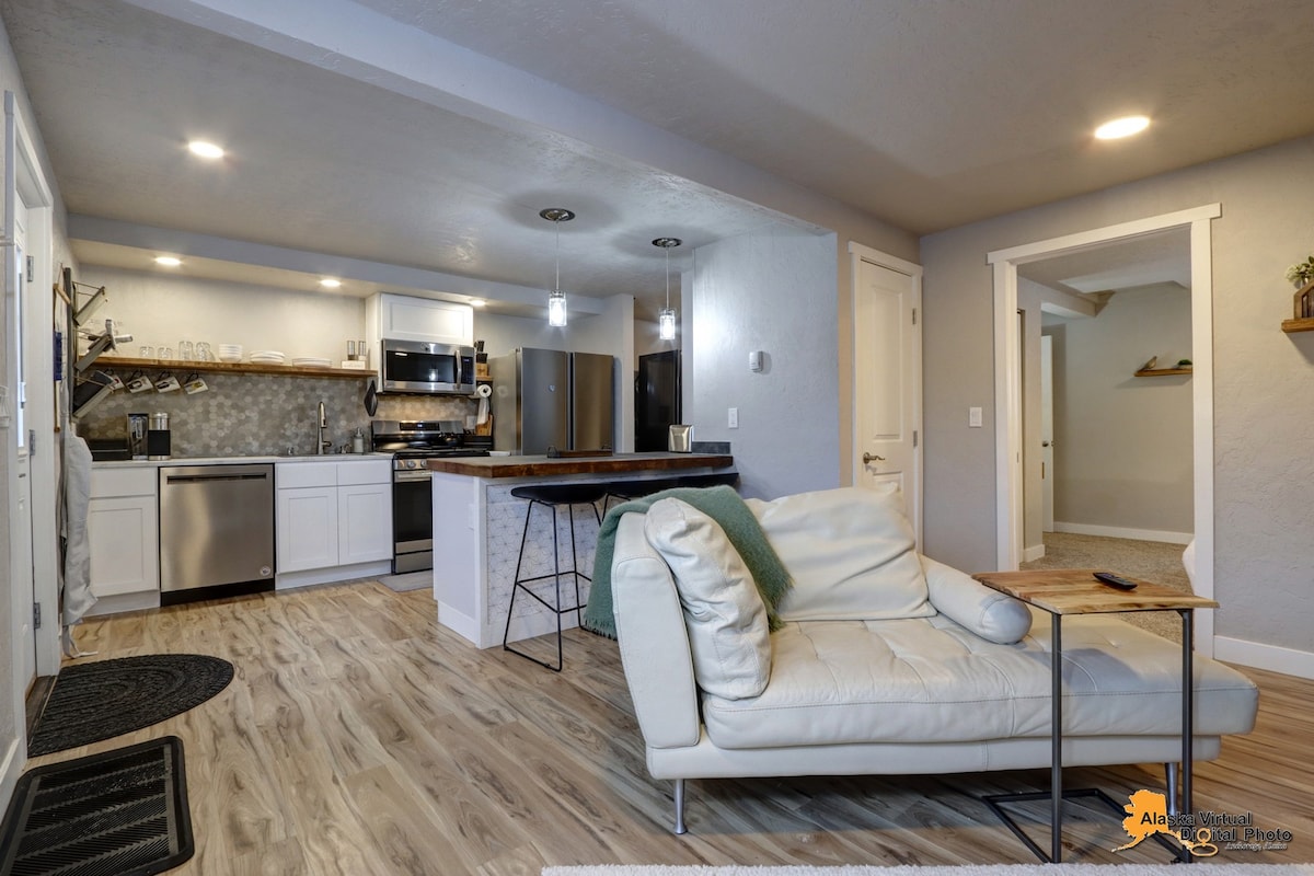 The image displays an open kitchen and living area with light wood flooring. A modern kitchen, equipped with stainless steel appliances and a unique bar-style seating area, is visible. A light-colored leather chaise lounge and a small side table are also present, creating a welcoming space.