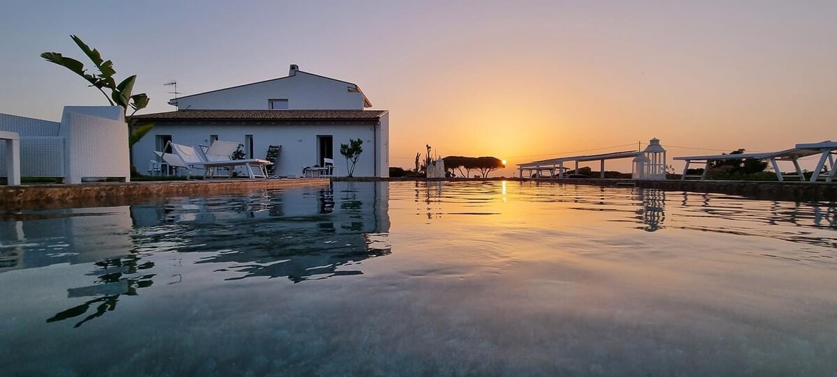 The image captures a tranquil pool reflecting the soft colors of a sunset. In the background, a white structure is visible alongside lounge chairs. A gentle silhouette of trees stretches across the horizon, contributing to the serene evening atmosphere.