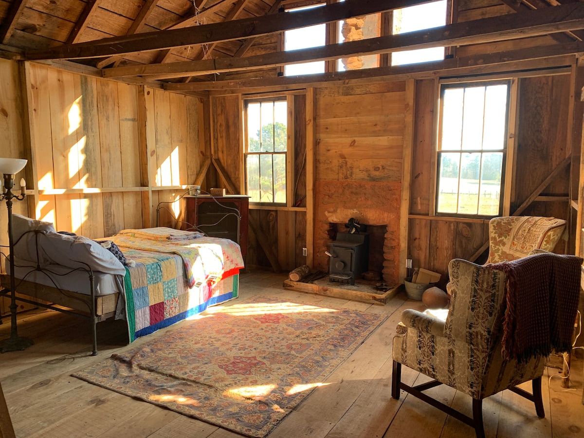 A rustic room features a bed with a colorful quilt, accompanied by a small stove and two windows allowing natural light to fill the space. A patterned rug is located underfoot, and a comfortable chair adds to the cozy setting.