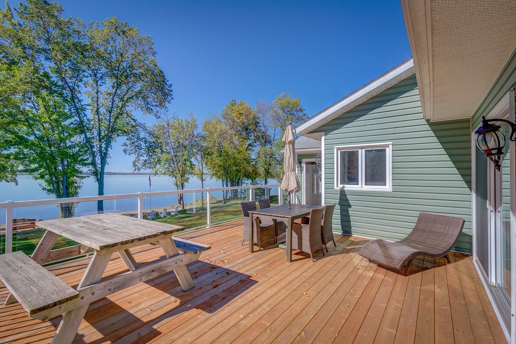 A spacious outdoor deck features a picnic table and dining set overlooking the water. A sun lounger is positioned nearby, surrounded by trees. The serene view captures the calm surface of the Ottawa River, enhancing the inviting outdoor space.