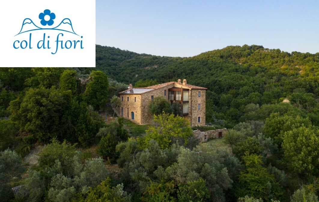 The exterior of Col di Fiori is shown, featuring a traditional stone structure surrounded by lush greenery and hills. The building has multiple levels with balconies and large windows, offering a harmonious blend with nature. The logo of Col di Fiori is displayed in the corner.