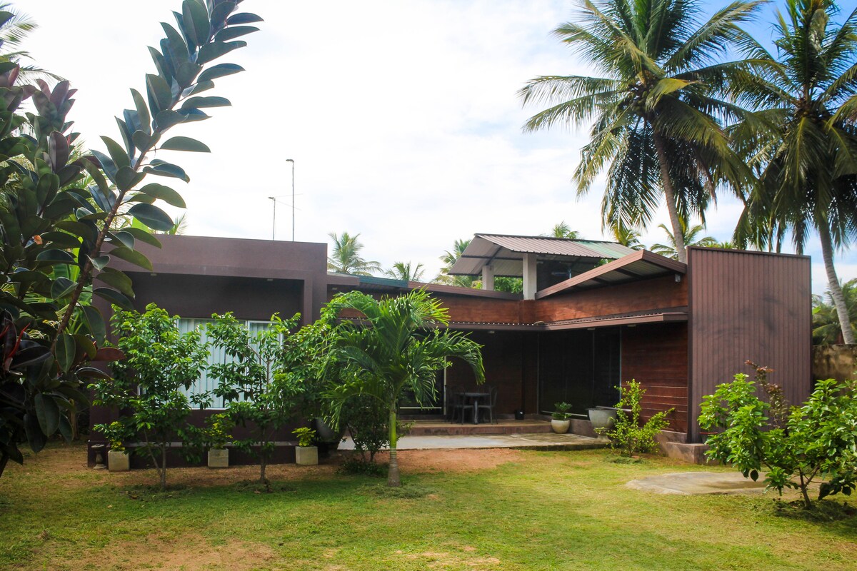 A modern container house is surrounded by lush greenery, featuring well-maintained palm trees and ornamental plants. A spacious garden area is visible, offering ample space for outdoor activities. The structure showcases a blend of wood and metal materials, enhancing the contemporary design.