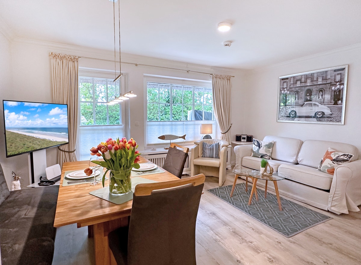 A light-filled living space includes a dining table set with flowers, a television, and a comfortable couch. Two large windows provide a view of greenery. Soft tones are present throughout the decor, emphasizing a relaxed atmosphere.