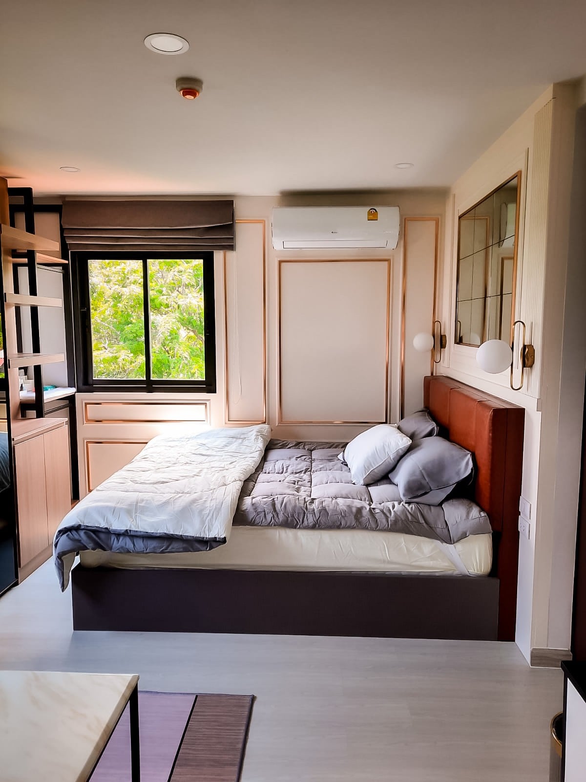 A well-lit bedroom is presented with a spacious bed dressed in neutral-toned bedding. A large window offers views of greenery, while light wooden accents and modern shelving enhance the room's design. Air conditioning is installed for comfort.
